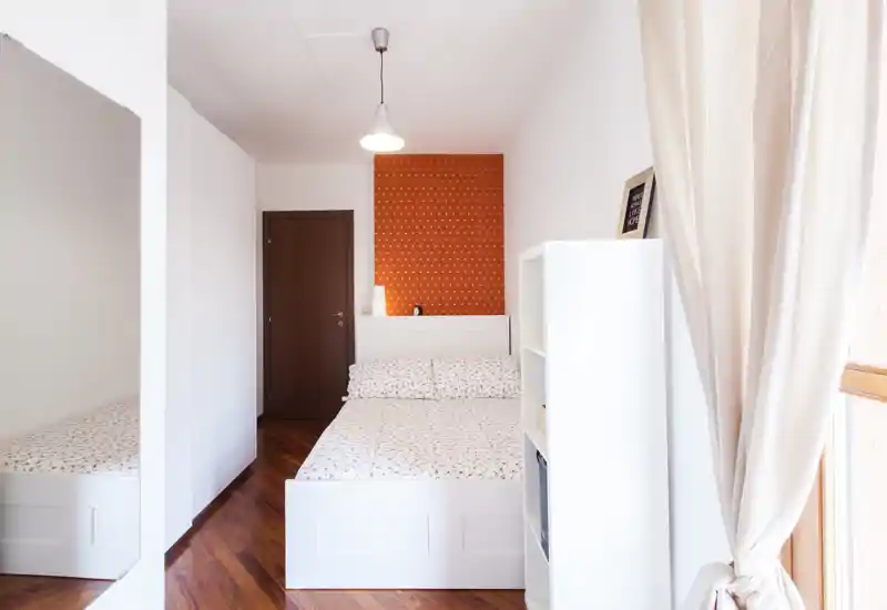 Dormitorio compacto y luminoso con cama blanca, ropa de cama estampada y una pared naranja de acento; la luz natural y el mobiliario limpio crean un ambiente acogedor.