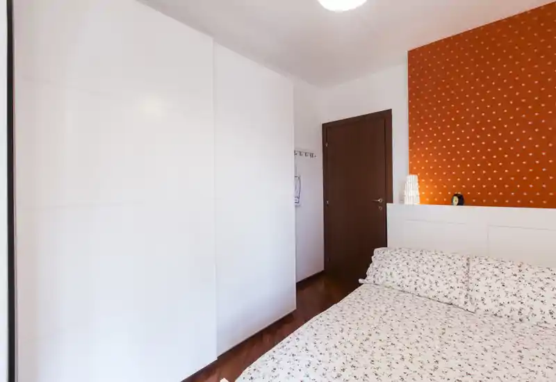 Dormitorio luminoso y acogedor con ropa de cama estampada, cabecero blanco y un gran armario empotrado blanco; pared naranja y lámpara de noche aportan calidez.