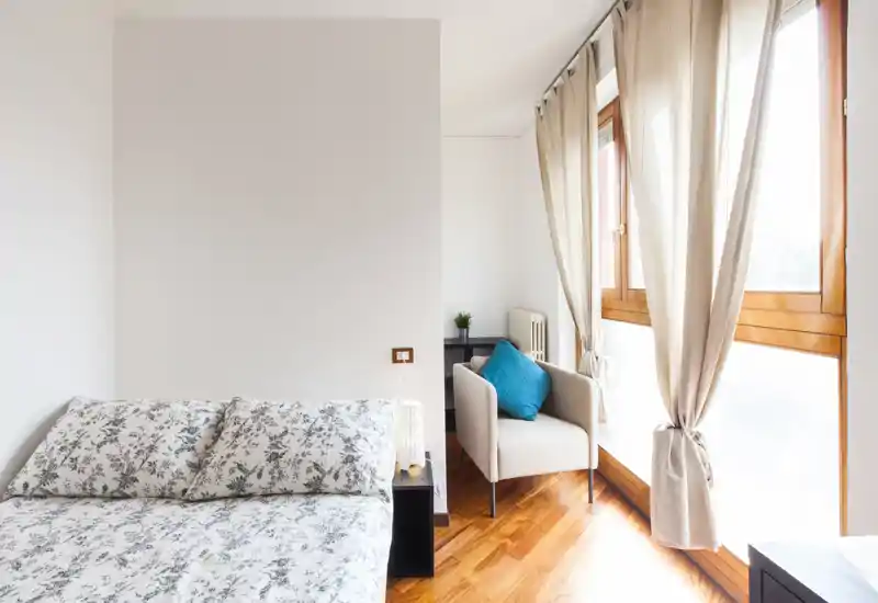 Dormitorio luminoso y aireado con ropa de cama estampada, un sillón cómodo con cojín turquesa junto a grandes ventanas y luz natural.