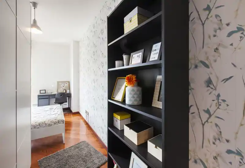 Dormitorio luminoso y bien decorado con cama individual, suelo de madera, estantería decorativa y un pequeño escritorio junto a la ventana — ideal para estudiante o persona sola.