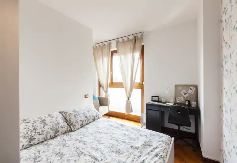 Dormitorio luminoso y bien decorado con cama doble con ropa de cama floral, un escritorio pequeño y un sillón junto a una gran ventana que aporta mucha luz natural.