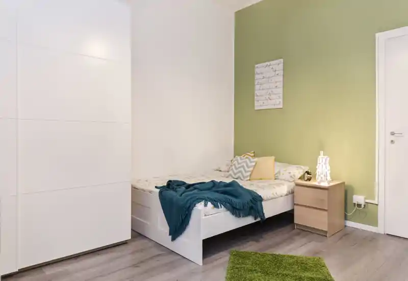 Chambre lumineuse et soignée avec un lit blanc, des coussins à motifs et un plaid bleu sarcelle contre un mur d'accent vert pâle. Une table de chevet en bois avec une lampe décorative et une armoire blanche complètent l'ensemble chaleureux.