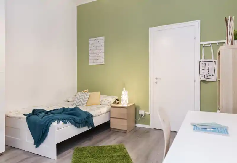 Chambre simple lumineuse et cosy avec lit blanc, table de chevet et bureau soigné ; textiles doux et lampe chaleureuse offrent une ambiance accueillante.