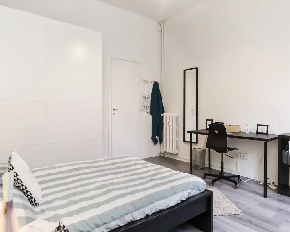 Dormitorio luminoso y minimalista con cama doble en primer plano, un gran armario blanco y un escritorio sencillo con espejo — ideal para una estancia cómoda o teletrabajo.