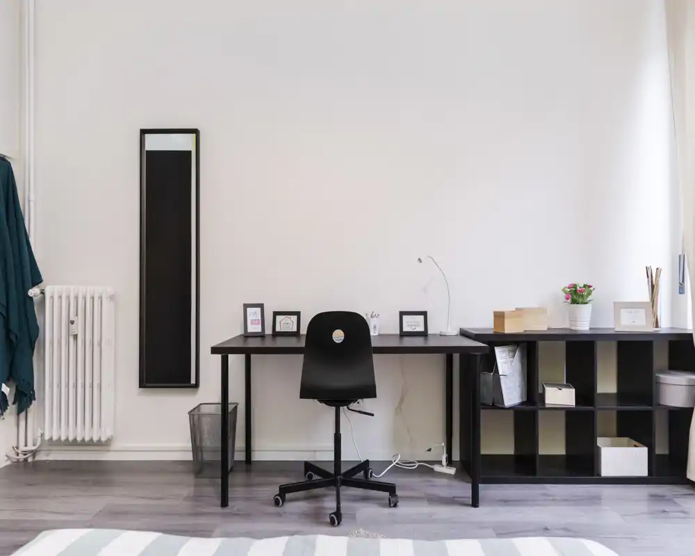 Rincón de dormitorio luminoso y minimalista con un escritorio largo y silla giratoria negra; estantería, marcos y una pequeña planta conforman un espacio de trabajo funcional.
