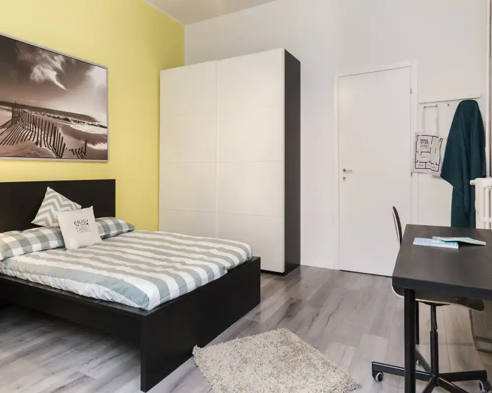 Dormitorio moderno y luminoso con cama doble, armario y escritorio; ropa de cama a rayas y una alfombra suave crean un ambiente acogedor y minimalista.