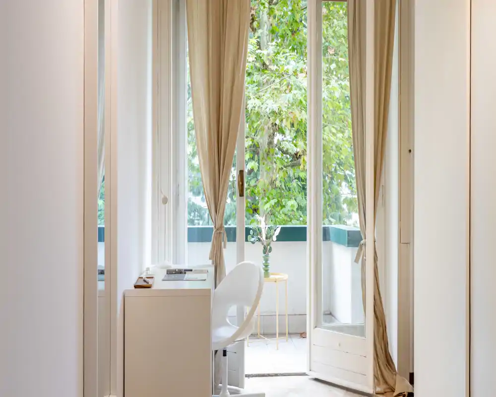 Small bright balcony seen through open French doors with neutral curtains, a compact desk and chair in the foreground and leafy greenery outside.