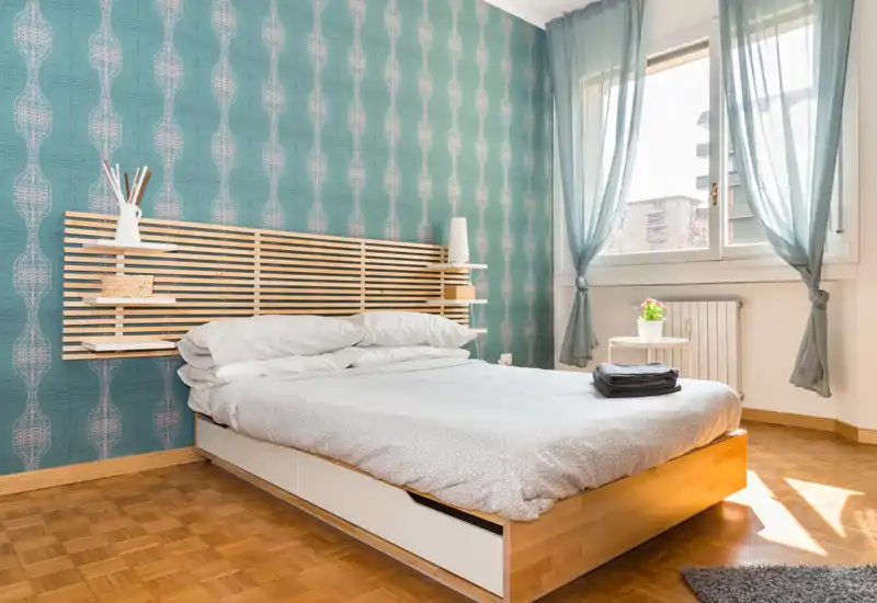 Camera da letto luminosa e moderna con un grande letto, parete decorativa turchese e luce naturale dalla finestra. Testiera in legno minimal e piccoli oggetti sul comodino rendono l’ambiente accogliente.