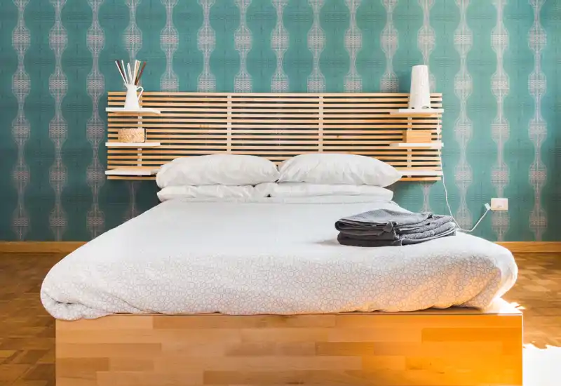 Camera da letto moderna e luminosa con un grande letto in legno, carta da parati turchese a motivo e biancheria piegata ordinatamente sul materasso. L'ambiente è pulito, accogliente e dallo stile minimale.