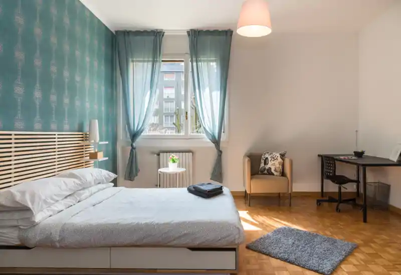 Camera da letto luminosa e di stile con letto matrimoniale, parete accentuata turchese e luce naturale; include una poltrona e una scrivania ordinata — perfetta per un soggiorno confortevole.