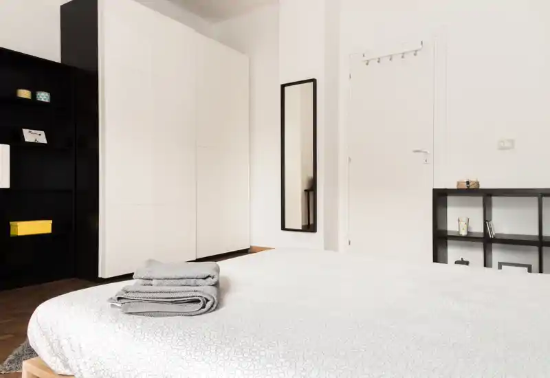 Camera da letto luminosa e minimal con letto ben rifatto in primo piano, grande armadio bianco, mensole nere e specchio a figura intera — ideale per annunci, mette in risalto ordine e spazio contenitivo.