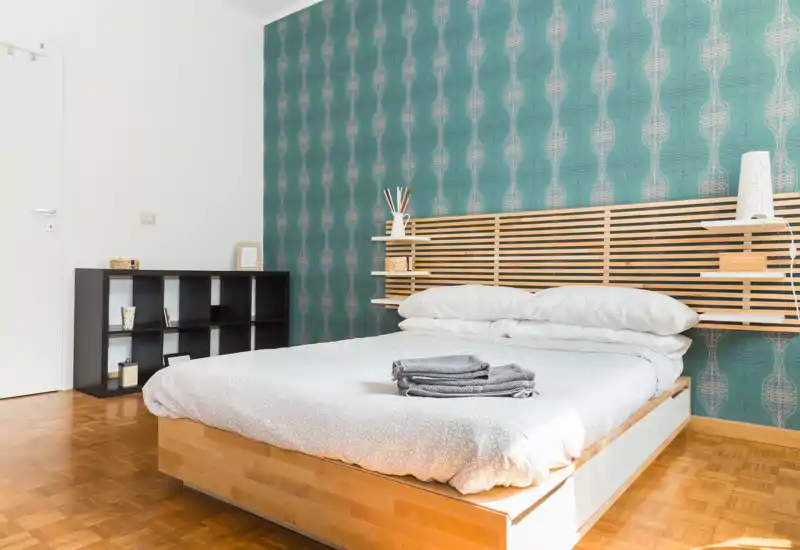 Camera da letto moderna e luminosa con letto a piattaforma in legno, parete decorativa turchese e mensole minimal; biancheria fresca e asciugamani piegati per un ambiente accogliente.