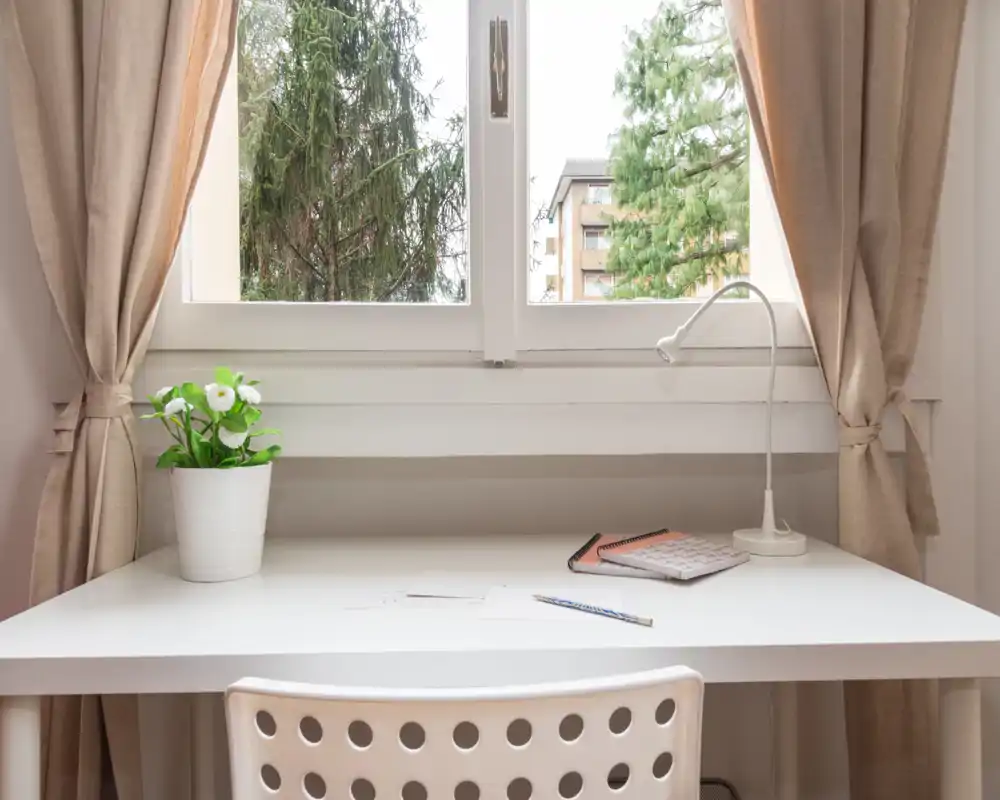 Espacio de trabajo luminoso y ordenado junto a la ventana con escritorio blanco, lámpara, cuaderno y planta decorativa — perfecto para trabajar o estudiar en un área de estar compacta.