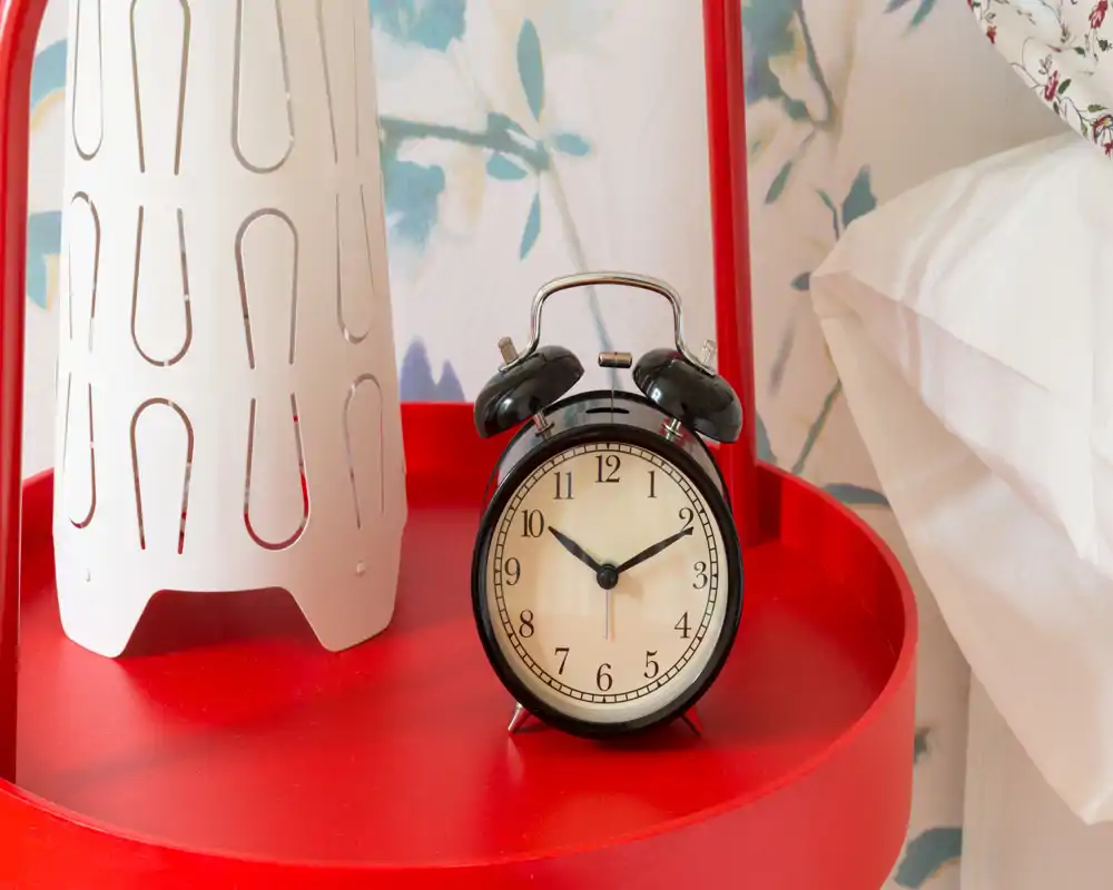 Plano detalle de una mesita de noche en un dormitorio con un despertador vintage, lámpara y un trozo de almohada con papel pintado — una imagen decorativa y acogedora.