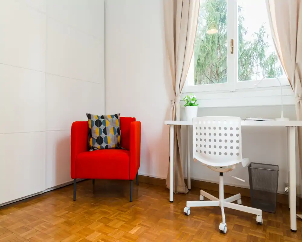 Zona de estar luminosa y minimalista con un sillón rojo como punto focal, cojín estampado y un pequeño escritorio junto a la ventana — ideal para un estudio o rincón de trabajo.