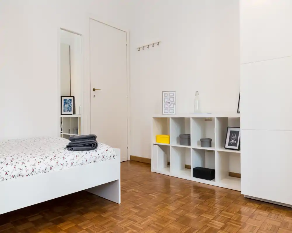 Dormitorio luminoso y minimalista con cama blanca de plataforma, ropa de cama estampada y toallas dobladas. Suelo de parquet, estantería de cubos con cajas decorativas y perchas en la pared que crean un espacio ordenado y acogedor.