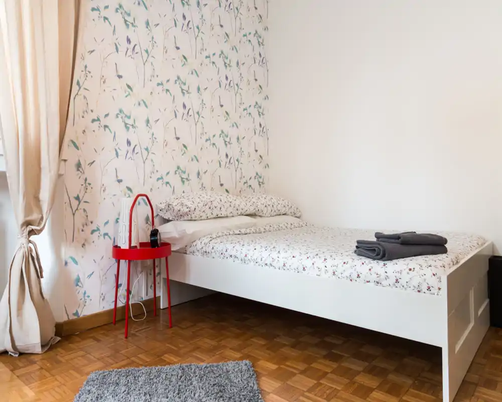 Dormitorio sencillo y luminoso con cama blanca, ropa de cama floral y luz natural suave. Una mesita roja y cortinas neutras completan el aspecto acogedor y moderno.