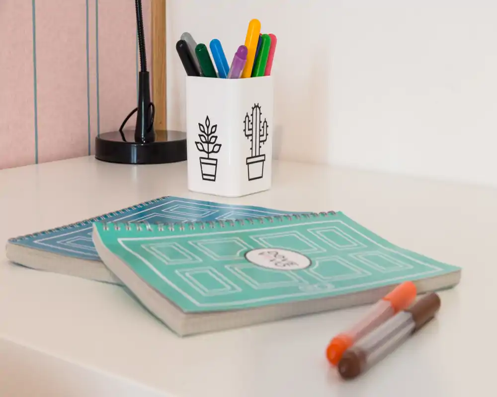 Primo piano di un angolo scrivania ordinato con due quaderni a spirale, penne colorate in un portapenne e una lampada su una superficie bianca — dettaglio di un piccolo spazio di studio/lavoro.