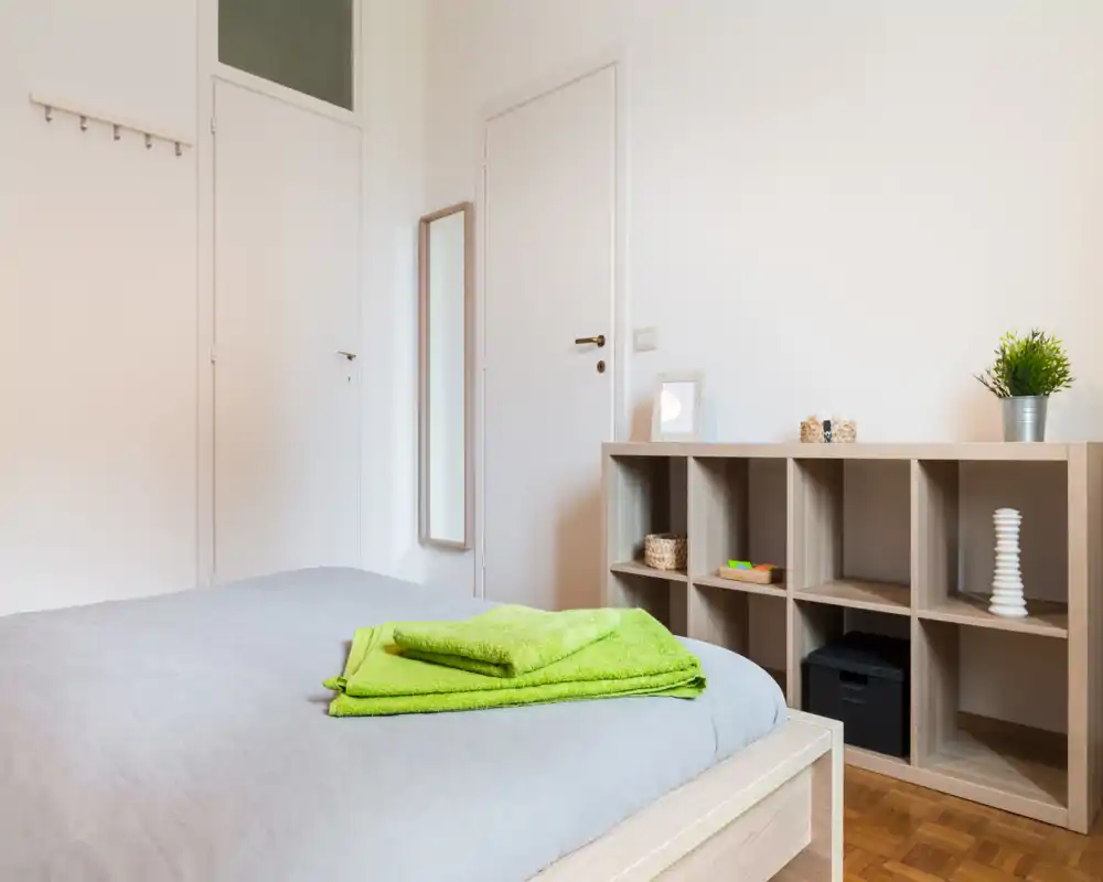 Camera da letto luminosa e minimale con letto in primo piano, scaffalatura in legno chiaro e semplici elementi decorativi; biancheria fresca e una pianta donano un tocco accogliente.