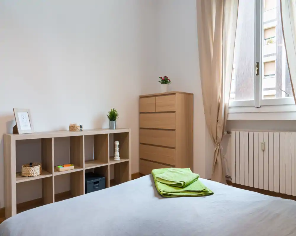 Camera da letto luminosa e minimal con letto ben rifatto, mobili in legno chiaro e luce naturale da una grande finestra.