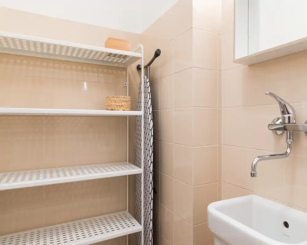 Baño compacto con un pequeño lavabo blanco, grifo cromado y paredes alicatadas; estantería y tabla de planchar visibles, espacio funcional y ordenado.