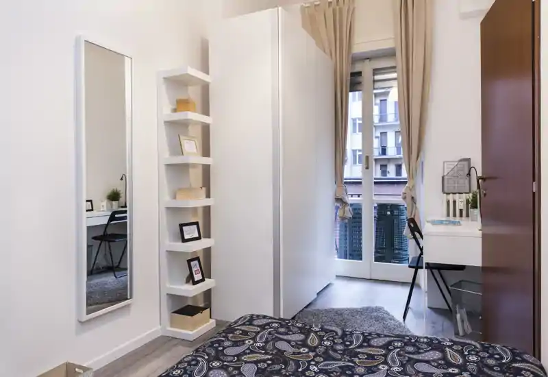 Dormitorio luminoso y ordenado con colcha estampada, espejo de cuerpo entero, estantería y escritorio junto a la puerta del balcón que aporta luz natural.