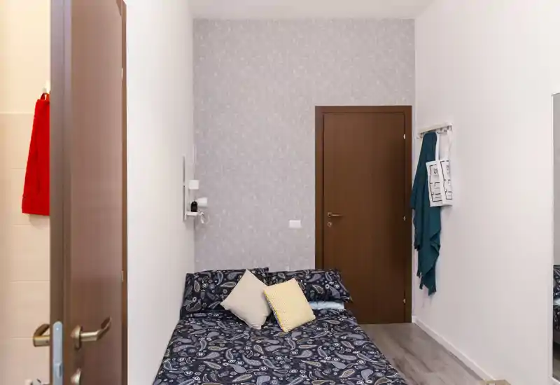 Dormitorio compacto con colcha estampada, cojines decorativos y papel pintado; luminoso, ordenado y adecuado para un anuncio de alquiler.