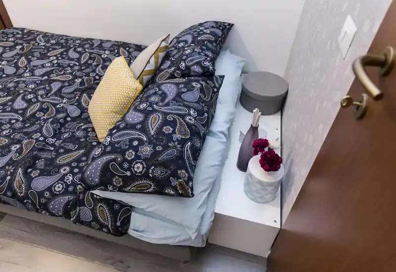 Dormitorio pequeño y estiloso con cama bien hecha, edredón estampado, cojines decorativos y mesita con accesorios — un espacio acogedor y bien presentado para un anuncio.