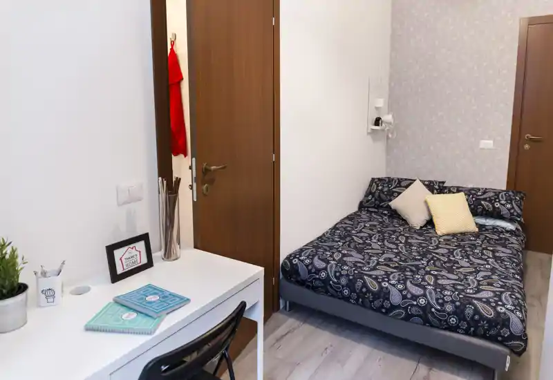 Dormitorio luminoso y bien arreglado con cama doble estampada, cojines decorativos y un escritorio compacto — ideal para una persona o pareja. Decoración moderna y buena iluminación que aportan calidez.