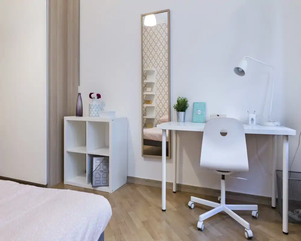 Rincón de dormitorio luminoso y ordenado con escritorio, silla y cama parcialmente visible; decoración minimalista y ropa de cama en tonos pastel que aporta tranquilidad.
