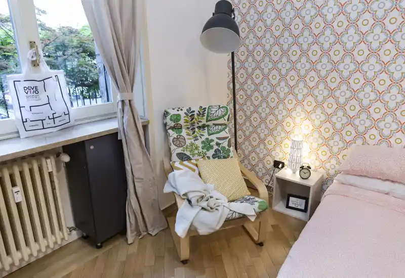 Rincón de dormitorio acogedor con pared decorativa, butaca con cojines y manta, mesita con lámpara y despertador, y cama parcialmente visible con ropa de cama rosa.