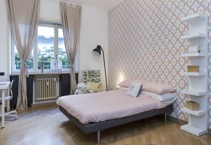 Dormitorio luminoso y bien decorado con cama doble, pared de diseño y una gran ventana que aporta luz natural. La habitación resulta acogedora gracias a la ropa de cama coordinada, la lámpara de pie y una zona de estudio junto a la ventana.