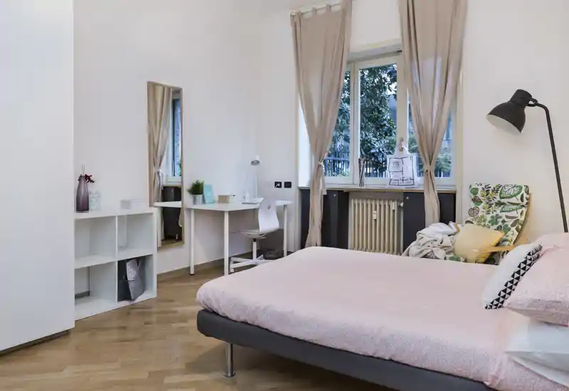 Dormitorio moderno y luminoso con cama doble vestida en rosa suave, un sillón acogedor con estampado, escritorio y gran ventana que aporta luz natural — ideal para estancias cómodas.