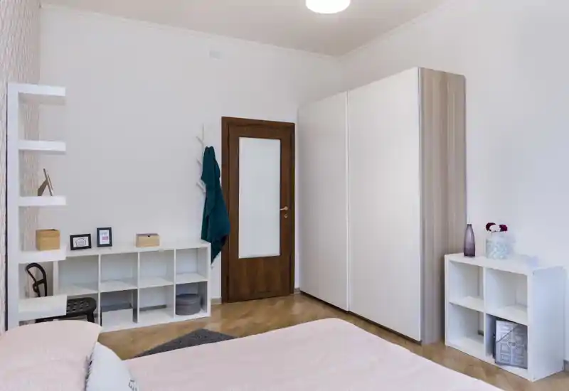 Dormitorio luminoso y ordenado con la cama parcialmente visible en primer plano, armario blanco grande, estanterías y suelo de madera clara: decoración moderna y serena, ideal para anuncios.