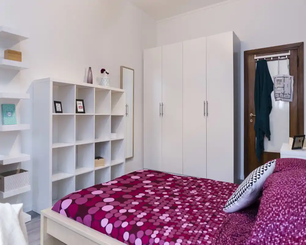 Dormitorio moderno y luminoso con colcha magenta estampada, armario blanco y estanterías. Líneas limpias, decoración sencilla y buena luz natural, perfecto para un anuncio de alquiler.
