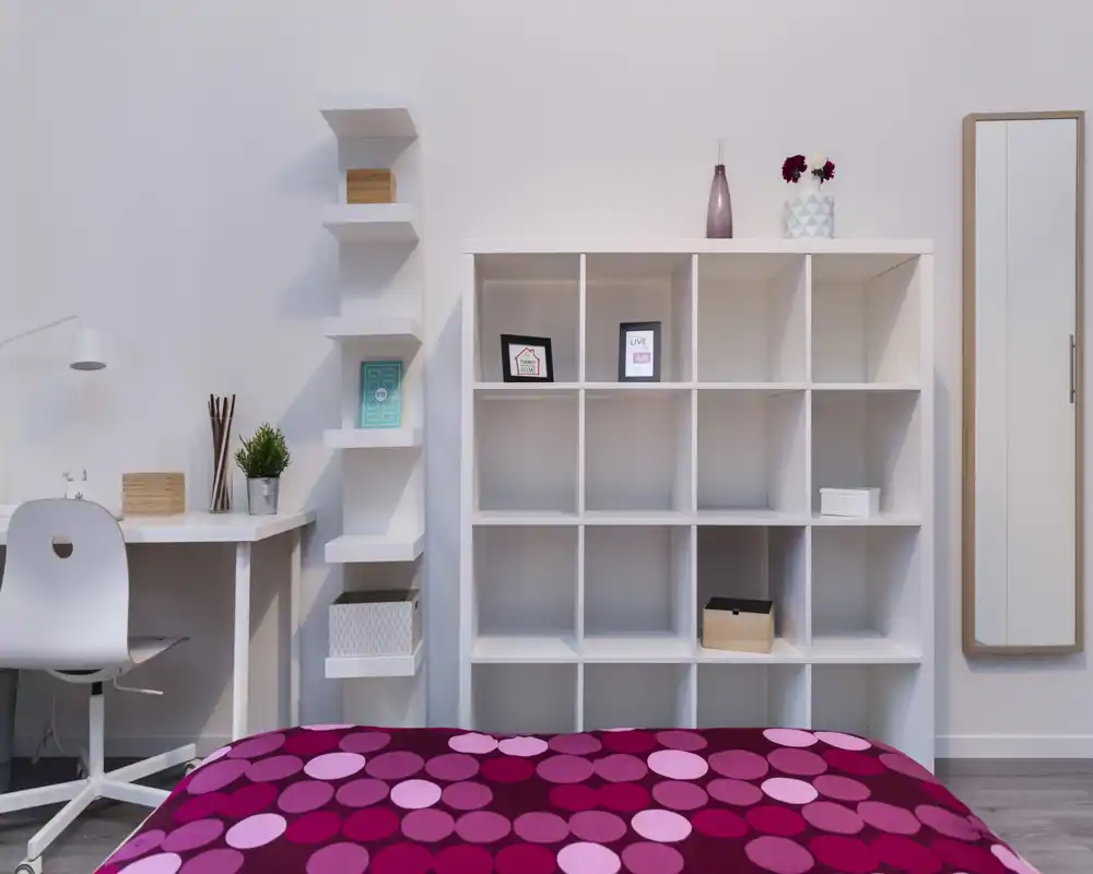 Dormitorio moderno y luminoso con colcha de lunares rosa y morado en primer plano, estantería blanca de cubos y escritorio minimalista; líneas limpias y detalles sencillos.