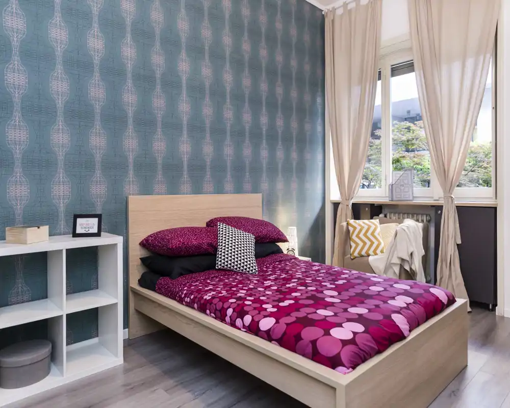 Dormitorio luminoso y con estilo con cama doble de madera, ropa de cama magenta con estampado, cojines decorativos y una gran ventana que aporta luz natural. Pared acento turquesa y estantería minimalista.