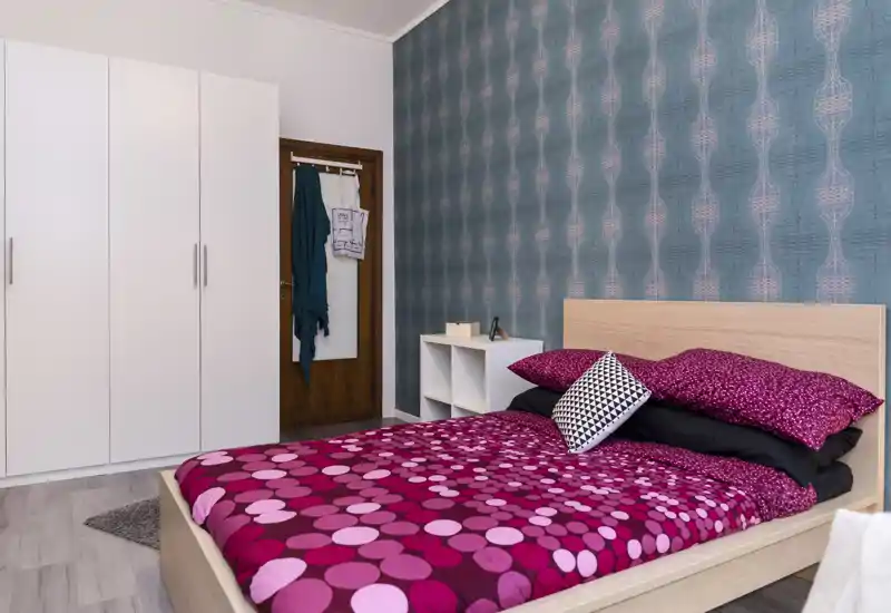 Dormitorio luminoso y bien presentado con cama de madera, ropa de cama estampada y papel pintado decorativo en turquesa; armario blanco y estantería junto a la cama.