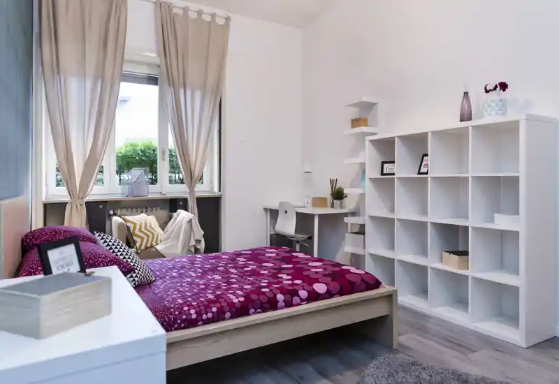 Dormitorio moderno y luminoso con edredón magenta estampado, almacenamiento junto a la cama y una zona de trabajo junto a la ventana. Espacio ordenado con cojines, estanterías y luz natural.
