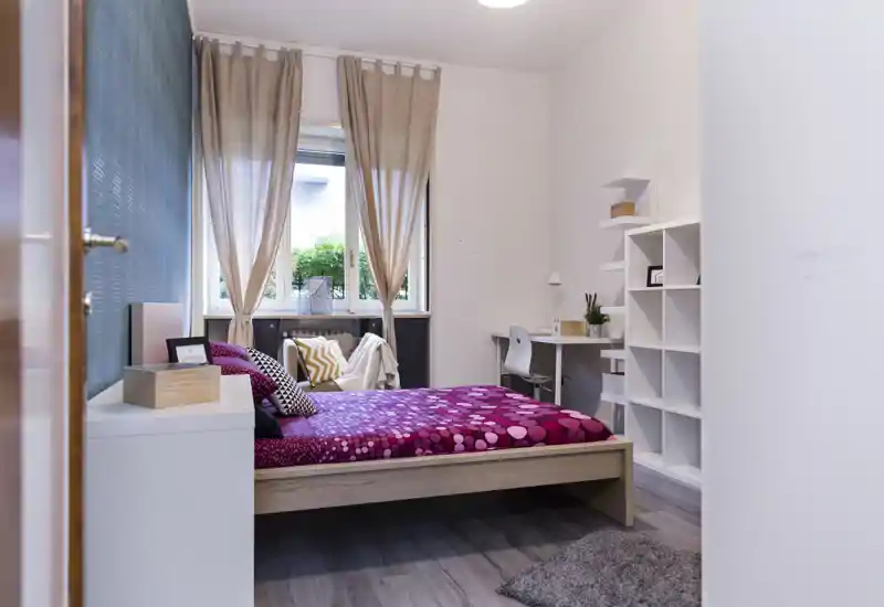 Dormitorio luminoso y bien decorado con cama doble con ropa de cama magenta estampada, cortinas suaves y un pequeño escritorio con estanterías — ideal para estudiante o una sola persona.