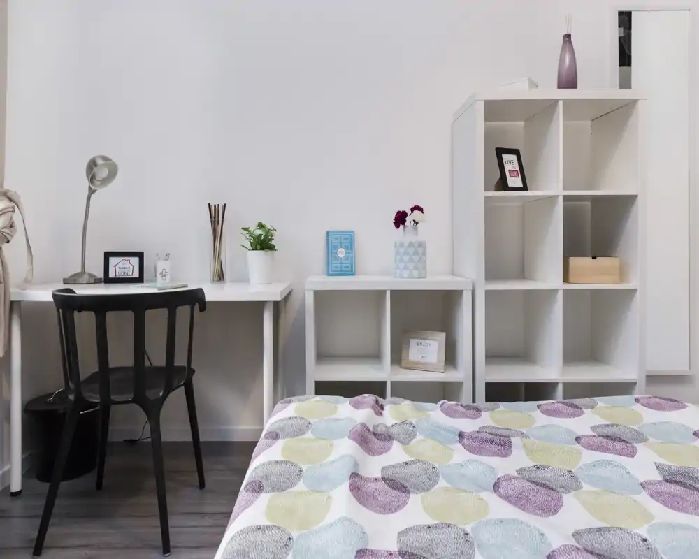 Chambre lumineuse et minimaliste avec édredon à motifs au premier plan, bureau simple et chaise, étagères ouvertes avec décorations — espace propre et bien éclairé parfait pour une annonce.