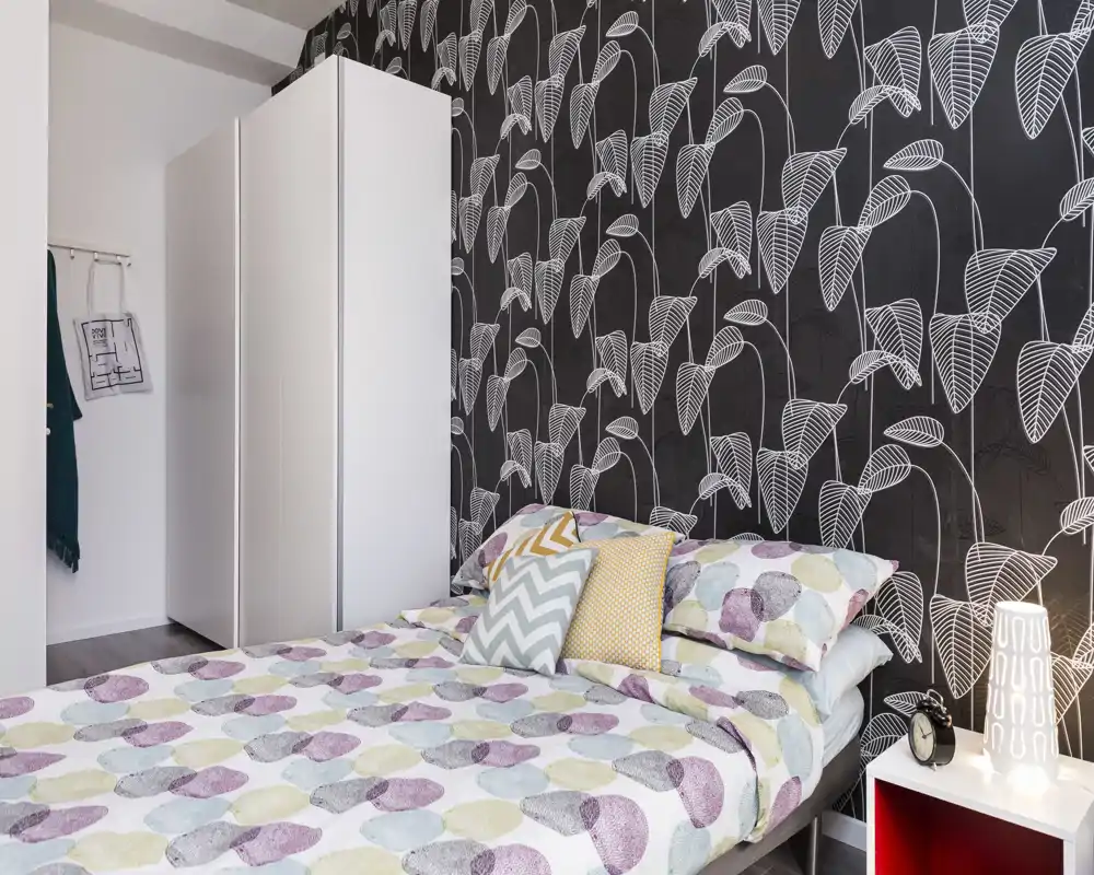 Chambre lumineuse et élégante avec papier peint à motifs et literie coordonnée ; lit double visible avec coussins décoratifs et lampe de chevet.
