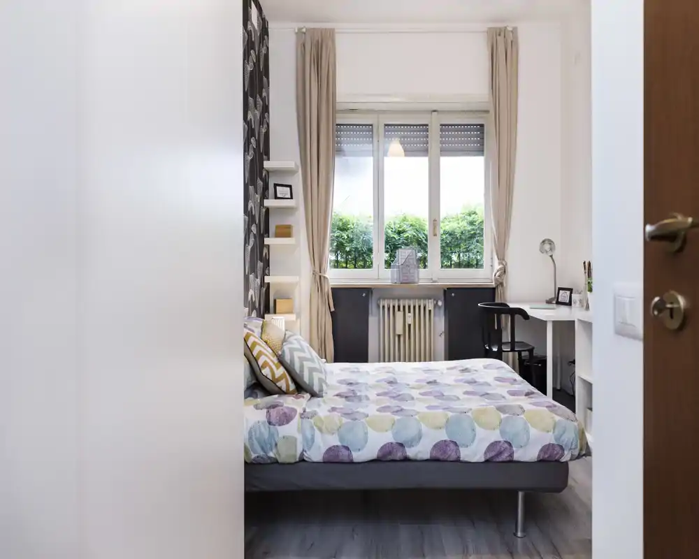 Chambre lumineuse et soignée avec un lit double à motif face à une fenêtre, rideaux clairs et un petit bureau à côté — parfaite pour une location compacte et confortable.