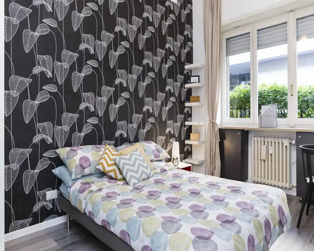 Chambre lumineuse et stylée avec papier peint à motifs, literie colorée et une grande fenêtre apportant de la lumière naturelle.