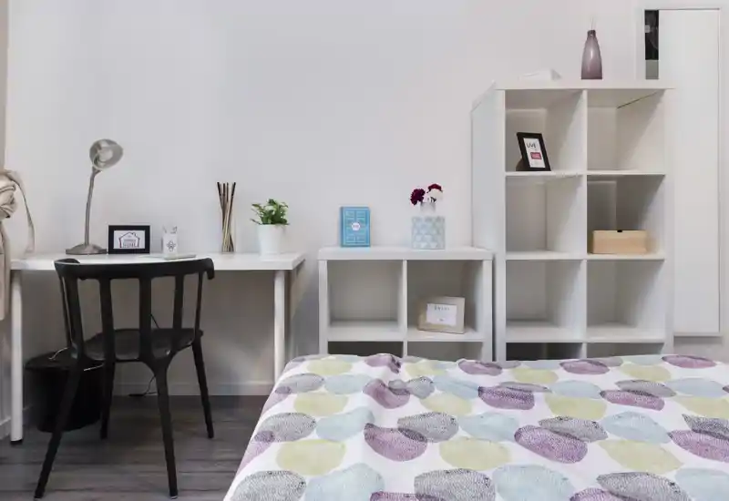 Chambre lumineuse et minimaliste avec édredon à motifs au premier plan, bureau simple et chaise, étagères ouvertes avec décorations — espace propre et bien éclairé parfait pour une annonce.