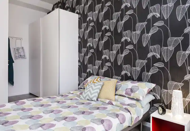 Chambre lumineuse et élégante avec papier peint à motifs et literie coordonnée ; lit double visible avec coussins décoratifs et lampe de chevet.