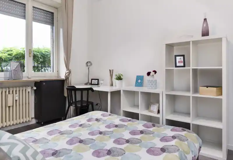 Chambre lumineuse et moderne avec couette à motifs, bureau près de la fenêtre et étagères ouvertes pour le rangement — idéale pour une personne ou courts séjours.