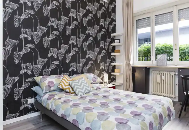 Chambre lumineuse et stylée avec papier peint à motifs, literie colorée et une grande fenêtre apportant de la lumière naturelle.