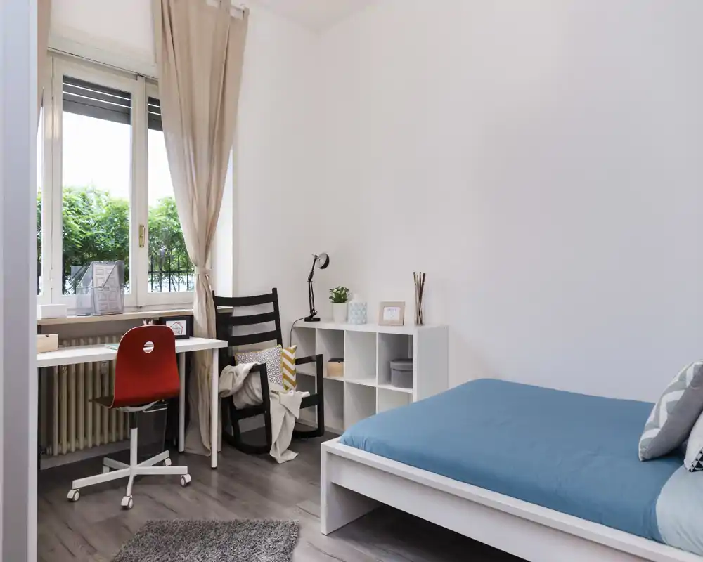 Bright, minimal bedroom with a single bed dressed in blue linen, a small shelving unit, and a work desk by the window letting in natural light.