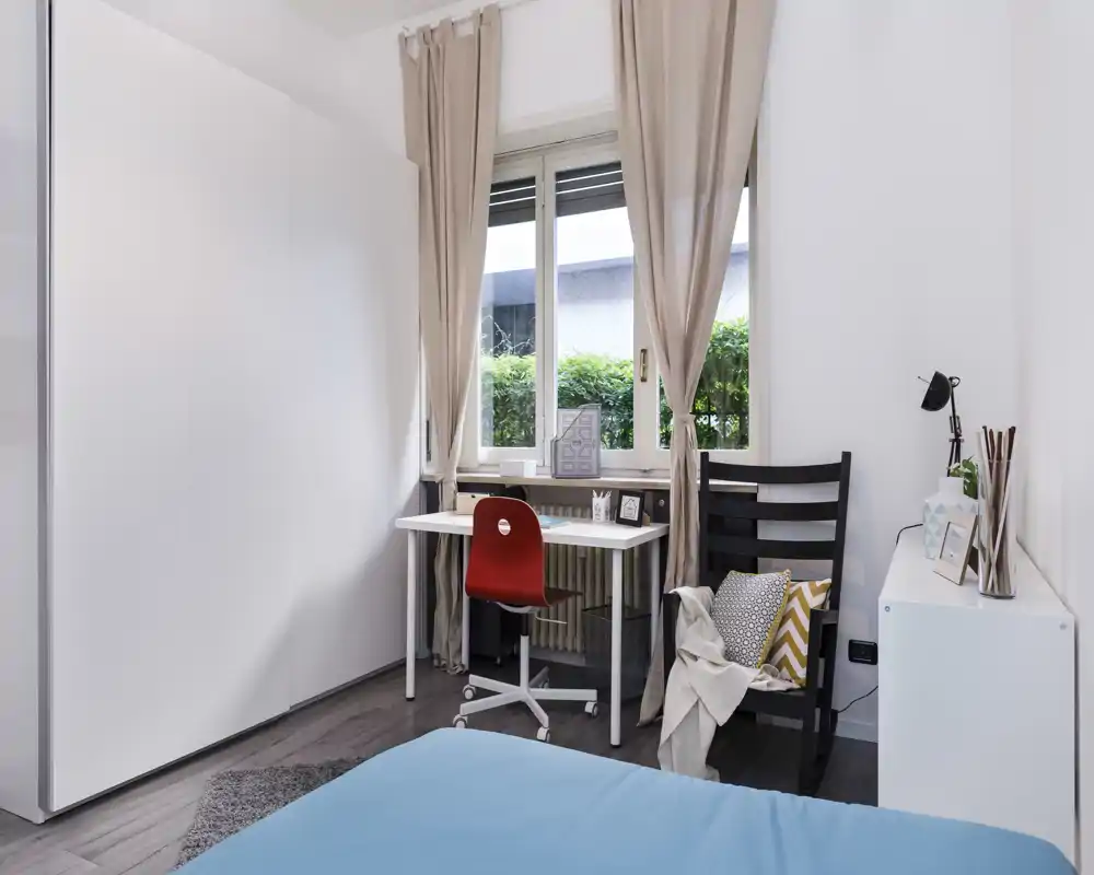 Bright, well-styled bedroom with a visible bed in the foreground, a neat desk by the window and cozy textiles that create a welcoming atmosphere.